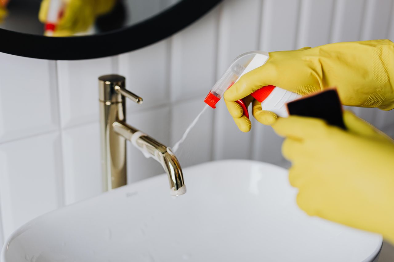 gallery-02 View from above of an unidentified housekeeper wearing yellow rubber gloves, spraying cleaning solution onto a shiny chrome bathroom faucet